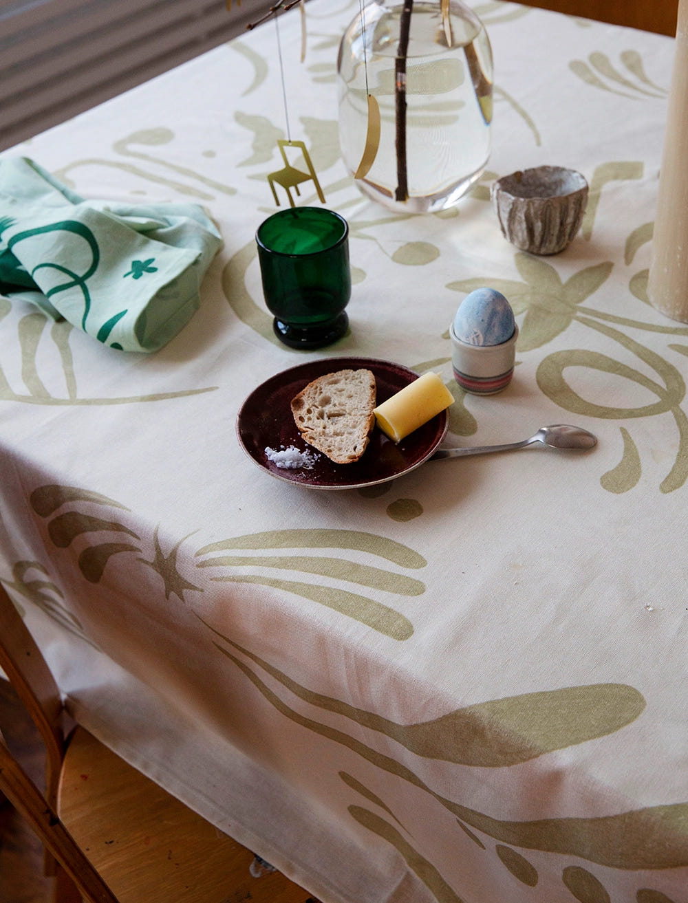 En vårig dukning på en beige mönstrad duk med en brödbit på ett fat, ett kokt ägg, ett grönt glas och en grönmönstrad servett ligger slarvigt bredvid.