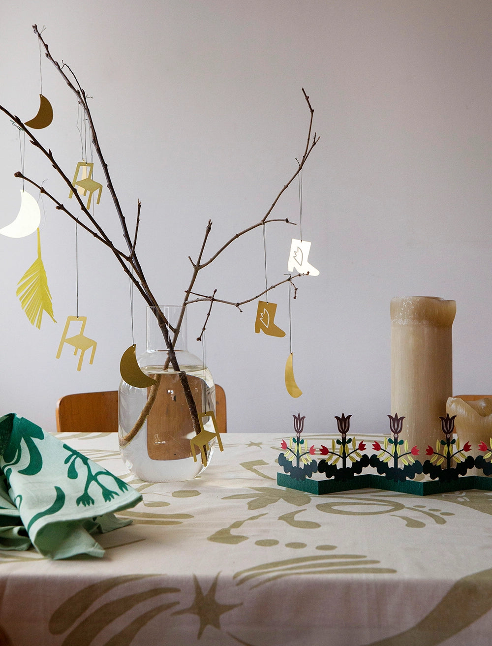 En vårig påskdukning med en beige mönstrad duk, en bordsgirlang i papper och en vas med några kvistar där hängen i form av boots, stol och måne i guldpläterat stål hänger.