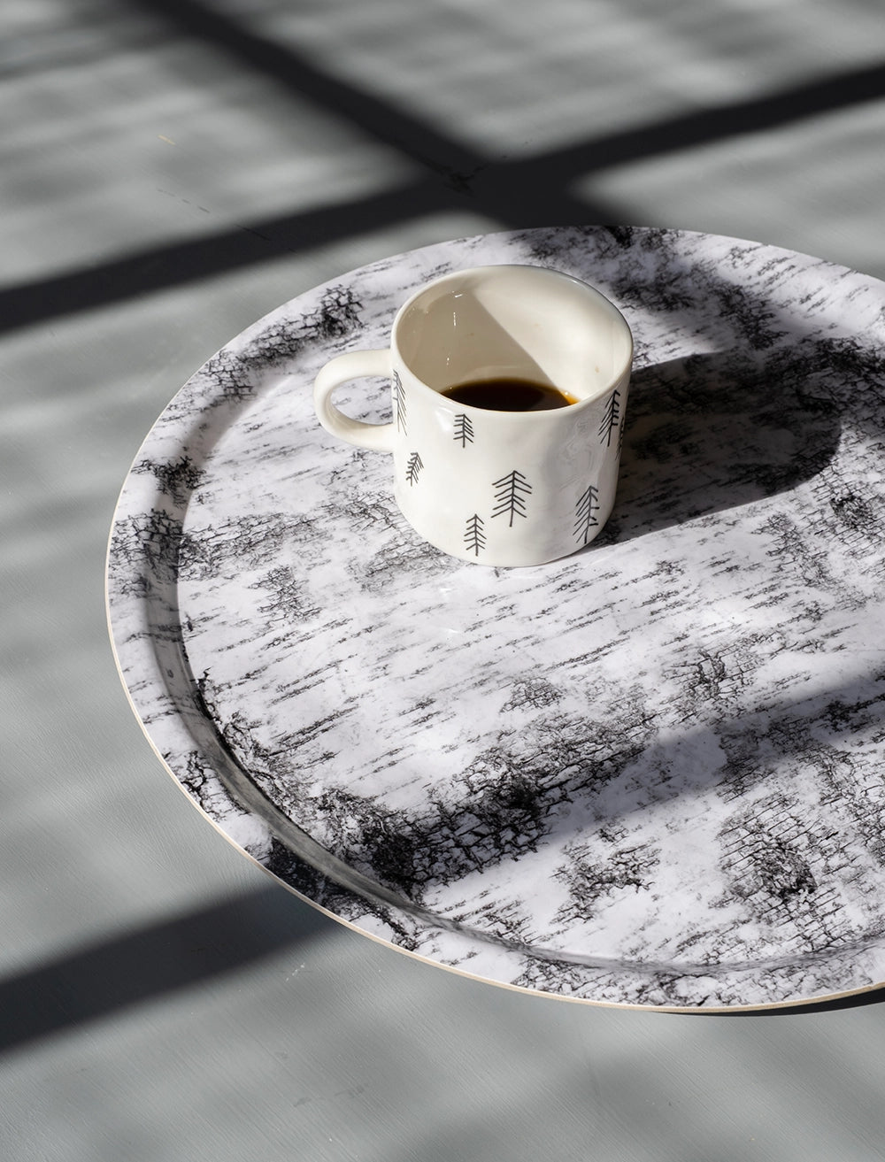 En bricka med näverbarkmotiv syns i solljus, på den står en vit porslinsmugg med kaffe i.