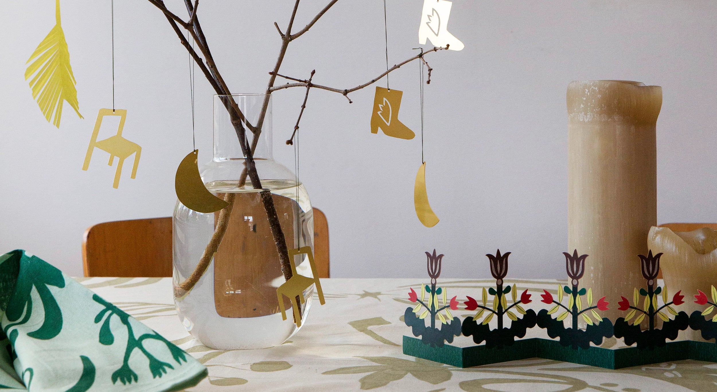 Ett dukat bord för påsk med en vas med kvistar och mässingshängen från FLD i form av stol, måne och boot, en bordsgirlang i papper, några blockljus. Allt på duken Högtid i beige.